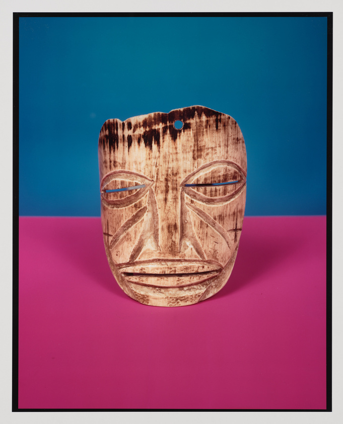 A carved wooden mask with tribal patterns displayed against a blue and pink background. The mask features prominent eyes, a nose, and a mouth, with a weathered texture and darkened areas around the edges and details, suggesting age or use. A small hole is visible at the top of the mask.