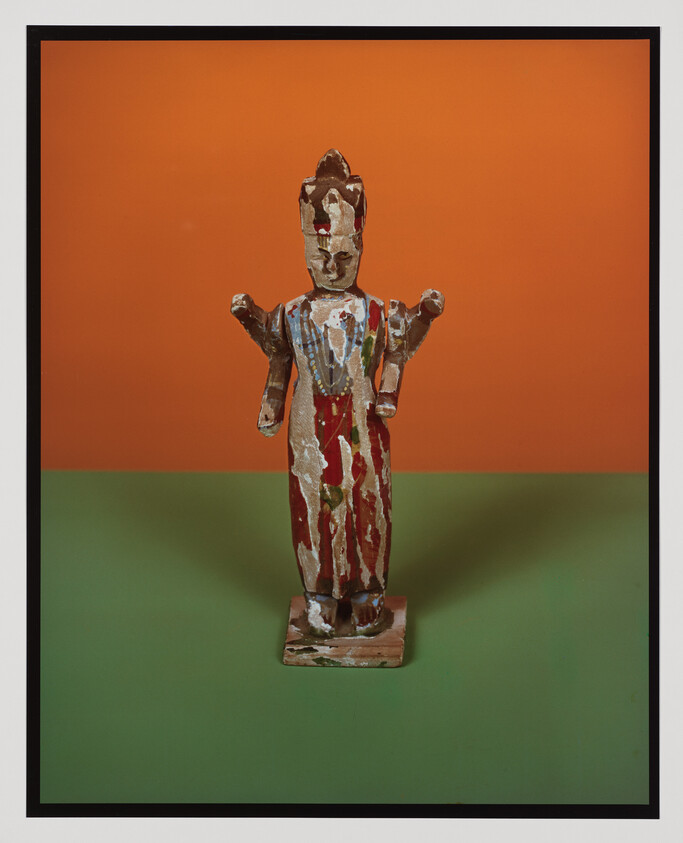 Worn painted wooden figurine with raised arms standing on a small base against orange and green background.