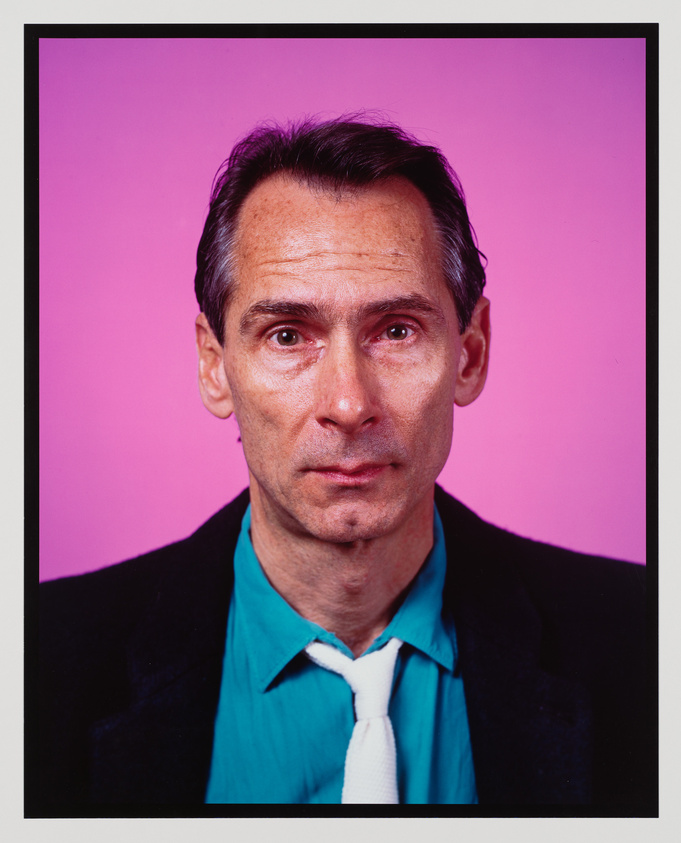 A portrait of a middle-aged man with prominent cheekbones and a thin face. He has short, neatly combed dark hair with streaks of gray and is gazing directly at the camera with a serious expression. The man is wearing a dark blazer over a bright teal shirt with a white tie. The background is a solid vibrant pink, providing a stark contrast to his attire.