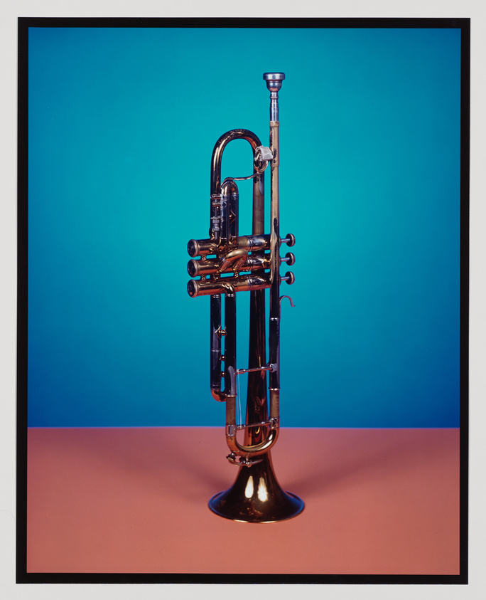 A brass trumpet with intricate valves stands upright against a blue background with a contrasting pinkish surface beneath it.