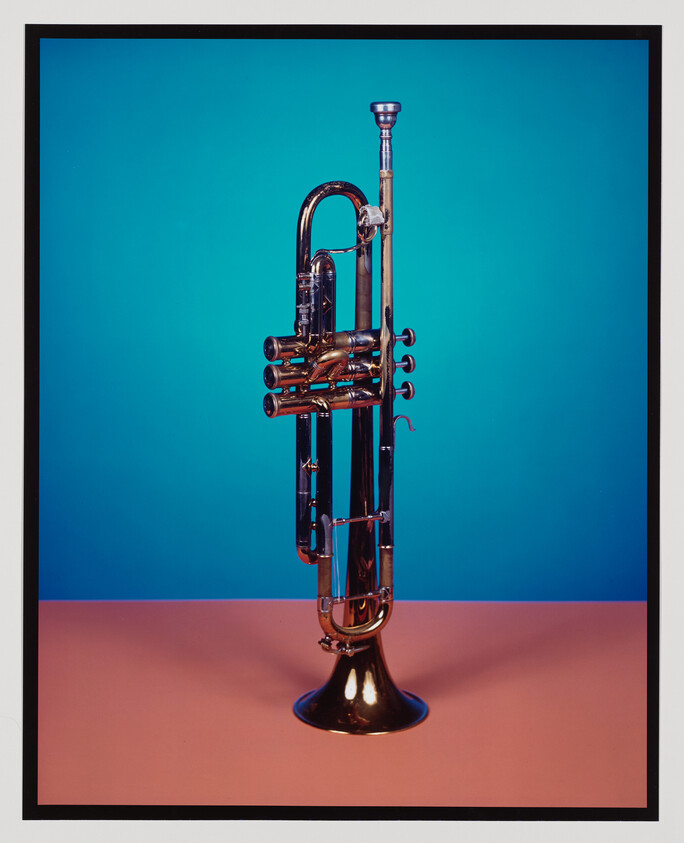 A polished brass trumpet standing upright on a pink surface against a bright blue background.