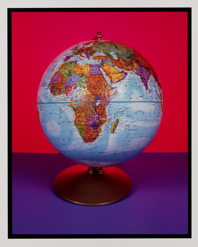 A colorful political world globe is centered against a vibrant red background with a purple surface beneath it, framed by a black border. The globe is tilted, showcasing continents in various shades, with countries and geographical details labeled.