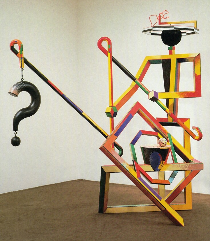 A colorful geometric sculpture with angled beams and a hanging black question-mark weight.