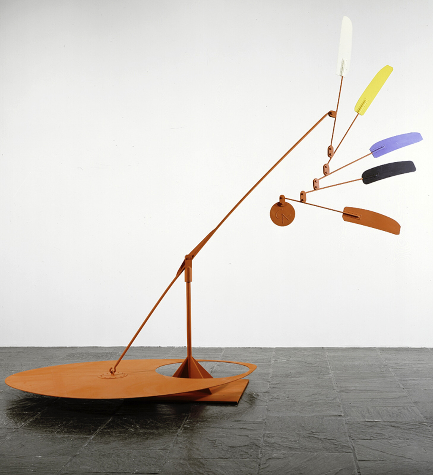 Orange metal balancing sculpture with long arm holding several colorful paddle-shaped weights in motion.