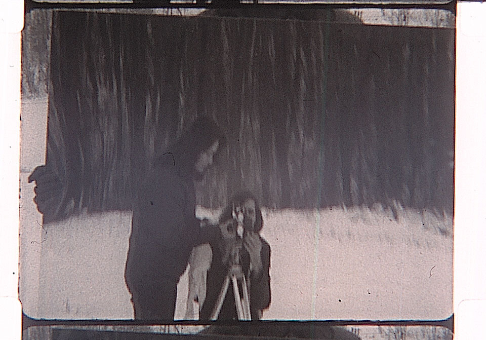 Two people set up an old camera on a tripod while standing on snowy ground.