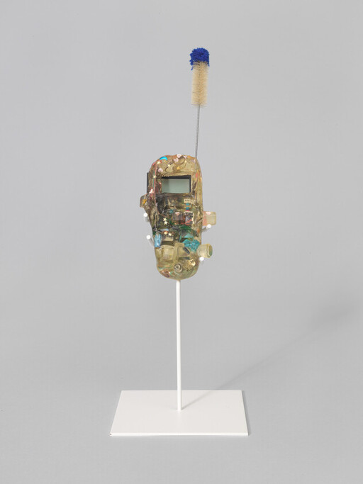 Decorated translucent mask-like sculpture on a white stand topped with a small brush.