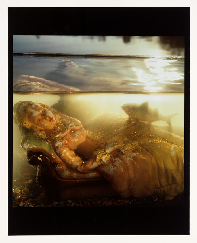 A reclining porcelain figurine on a chaise lounge lies submerged underwater with a fish swimming nearby.