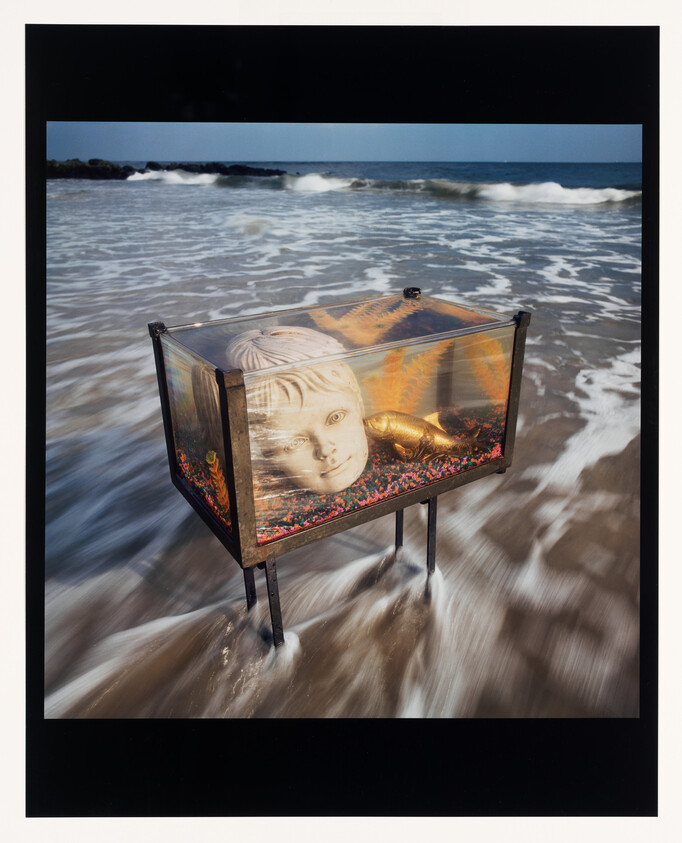 A glass tank on legs holds a large sculpted child head and gold figure standing in ocean surf.