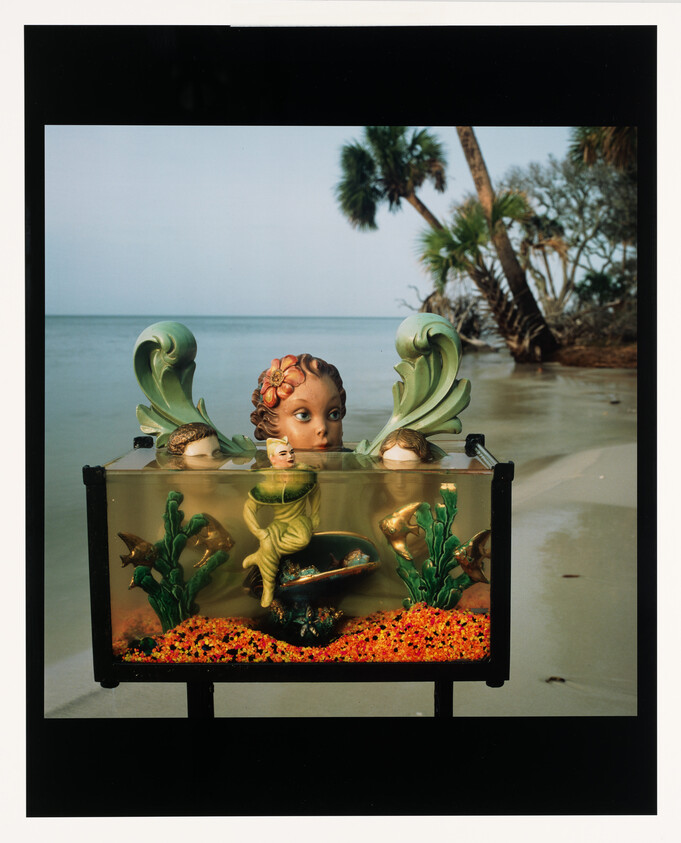 A decorative aquarium with a sculpted child's head and mermaid figure standing on a sandy beach.
