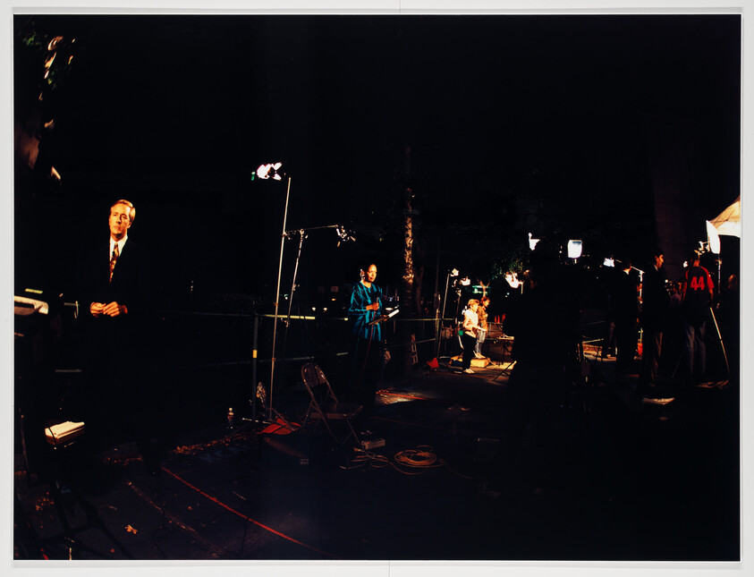 News crew sets up lighting and cameras at night while reporters prepare to deliver live segments.