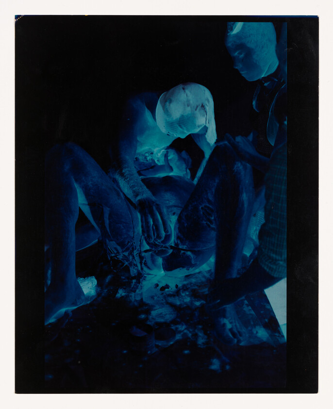 A cyanotype photograph depicting two individuals engaged in an activity, with one person sitting and the other leaning over, both appearing to focus on an object or task at hand. The blue-toned image creates a surreal and monochromatic effect.