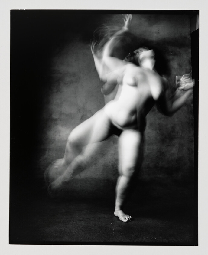 Blurred nude figure captured mid-dance with flowing, overlapping movement against a dark background.