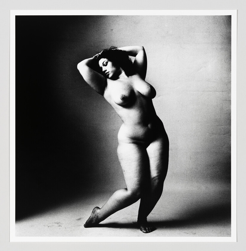 Nude woman standing on tiptoe with crossed legs and hands behind her head, gazing downward.