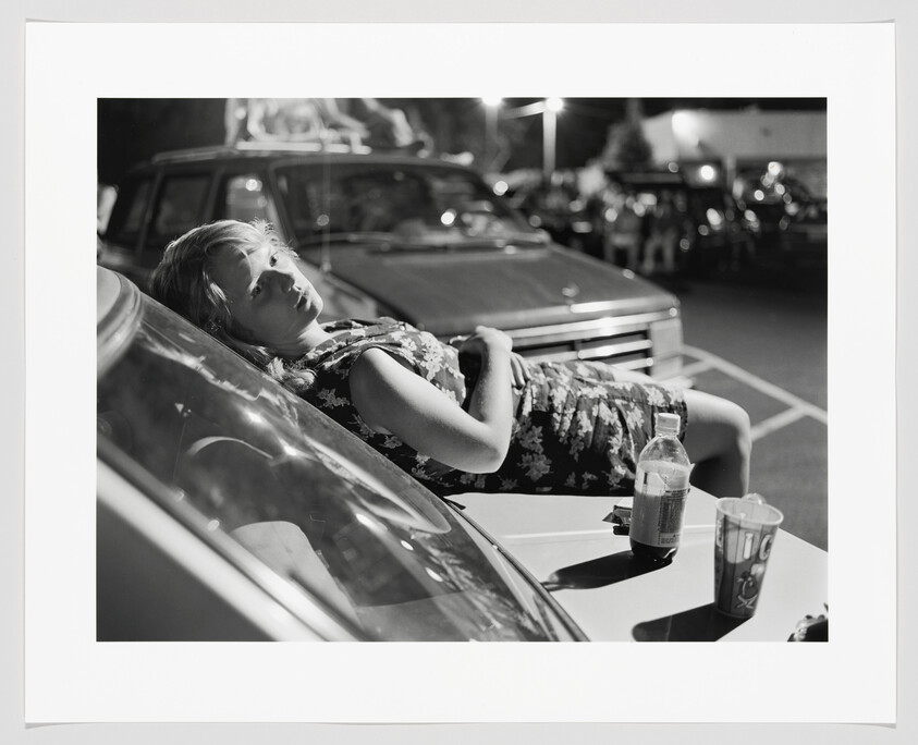 A woman in a floral dress lies on a car hood in a nighttime parking lot.