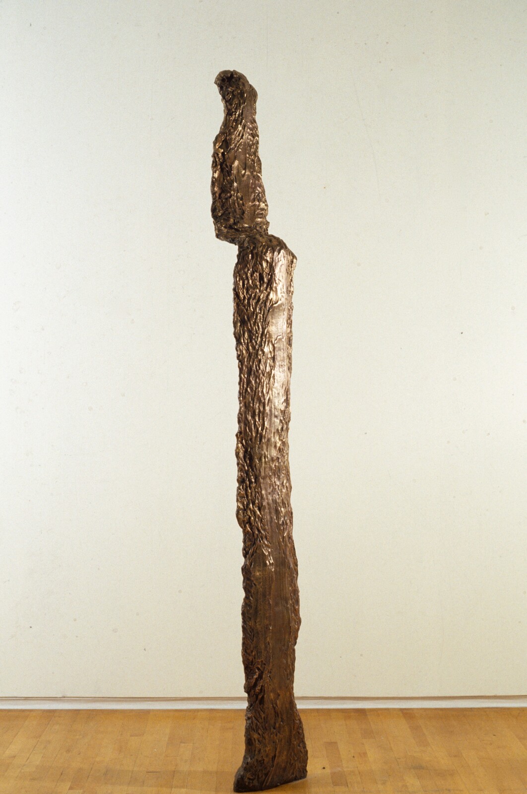 Tall thin bronze sculpture resembling a standing human figure with rough texture on a wooden floor.