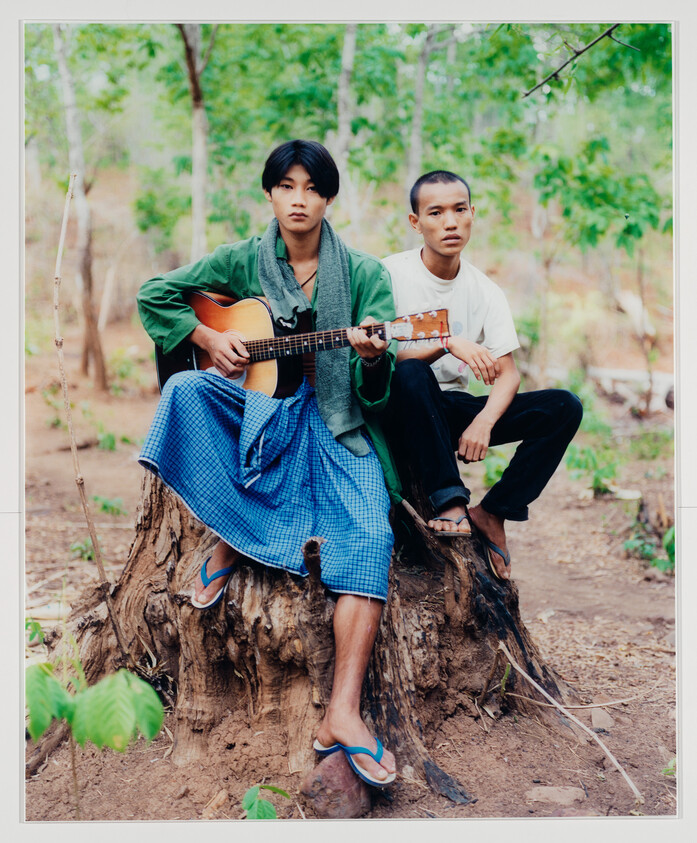 Two young men sit on a tree stump in a forest, one playing an acoustic guitar.