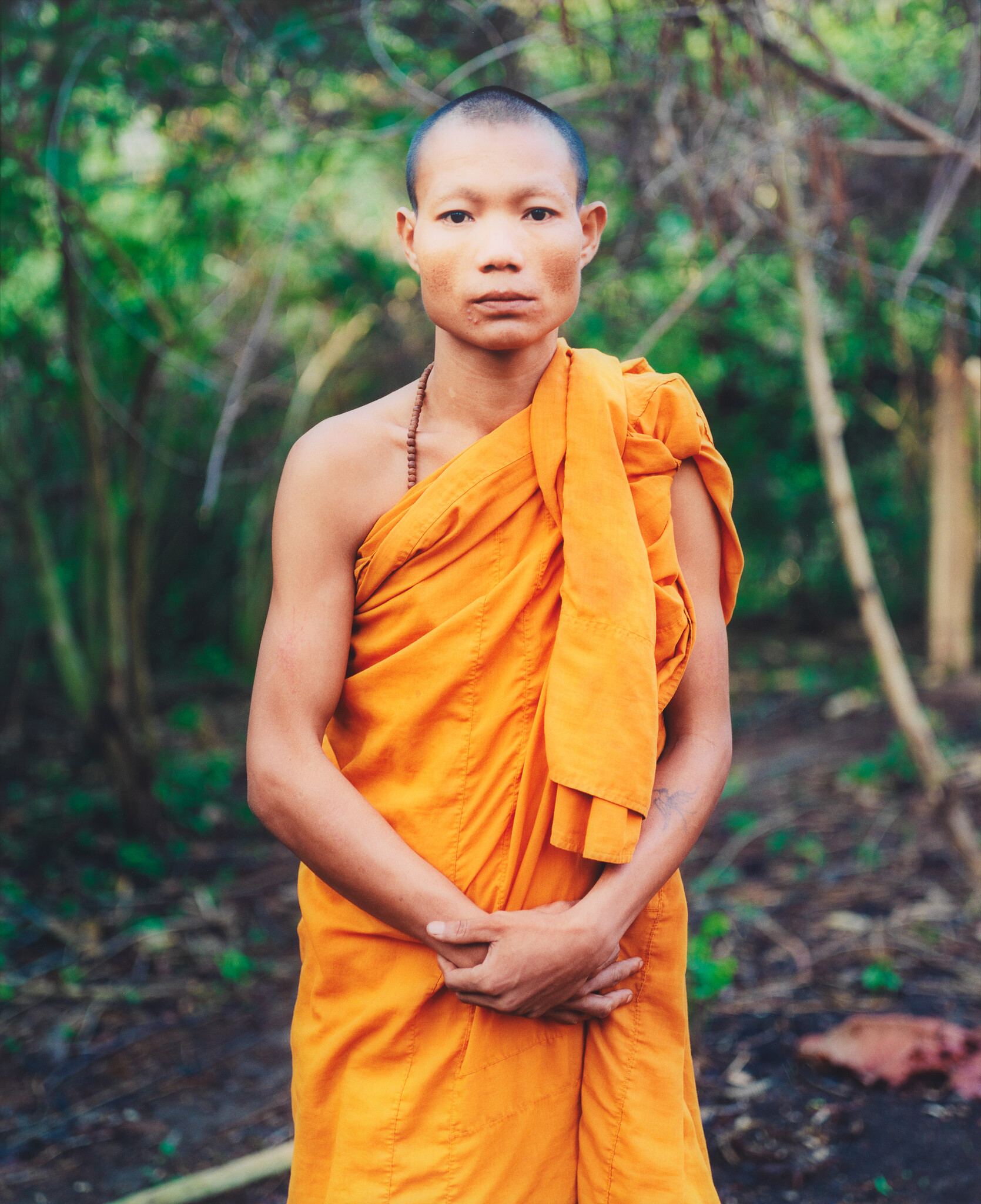 Young Buddhist monk stands in orange robe with hands clasped in a forest setting.