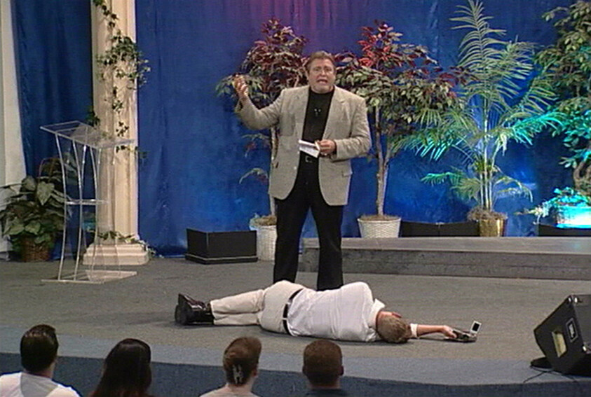 Man speaks on stage while another person lies facedown on the floor reaching toward a phone.
