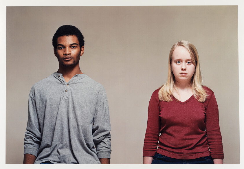 Two young adults standing side by side against a neutral background, looking directly at the camera with neutral expressions.
