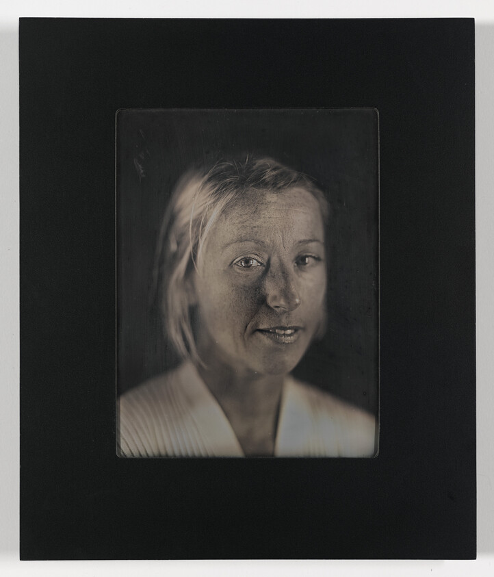 A monochromatic portrait of a woman with her hair pulled back, centered on a dark background, with a contemplative expression. The image has a vintage feel, possibly created using an old photographic process, and is framed within a black border.