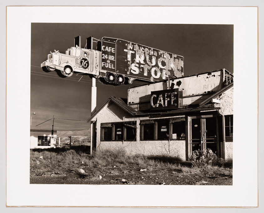 A black and white photograph depicting an old, dilapidated truck stop with a large, vintage billboard above it. The billboard features a three-dimensional truck and trailer model and advertises a 24-hour cafe and fuel. The words "WELCOME TRUCK STOP" are visible on the sign, which towers over the abandoned building below. The surrounding landscape is barren and the sky is clear.