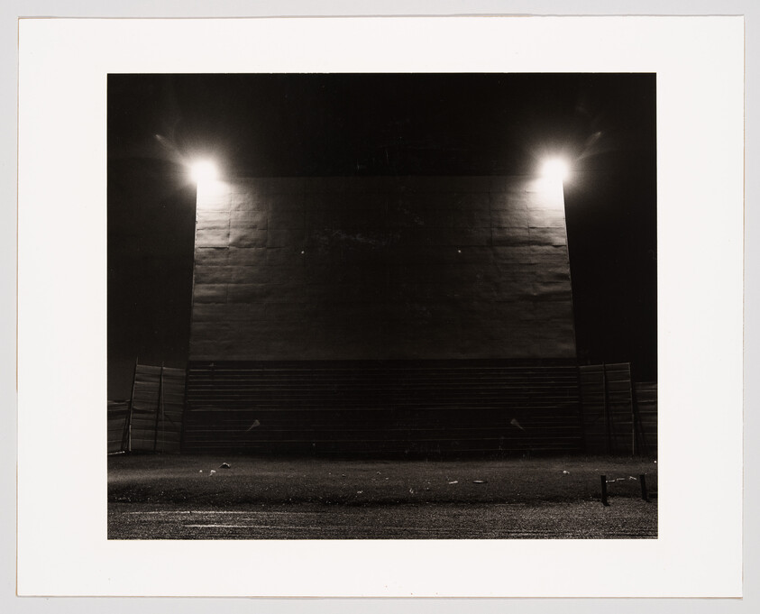 A black and white photograph depicting a large, dark wall illuminated by two bright lights at the top, casting a glow on the wall's surface. In the foreground, there is an empty, flat area with a few small objects scattered on the ground, and a fence partially visible on the left side. The overall scene is nocturnal and desolate.