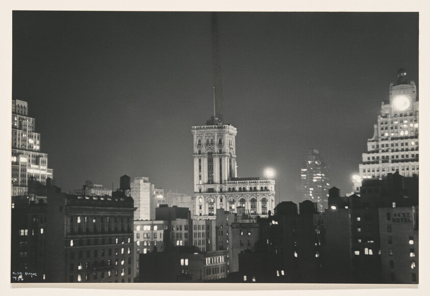 Tall ornate tower lit at night rises above surrounding city buildings.
