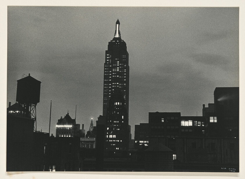Empire State Building silhouette rises above dark Midtown Manhattan skyline with lit windows.