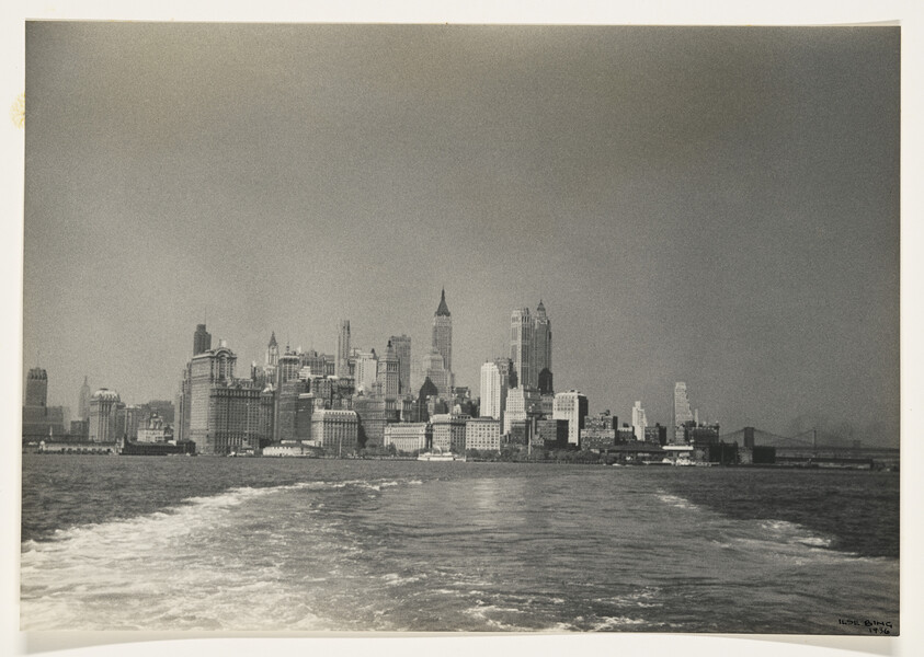 A vintage black and white photograph of the New York City skyline viewed from the water, with a clear view of several iconic skyscrapers and a bridge faintly visible in the background. The water in the foreground shows ripples and waves, suggesting the photo was taken from a moving boat.