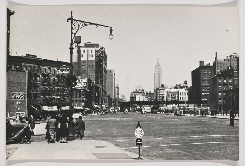 People waiting near a bus stop on a wide city street with the Empire State Building visible.