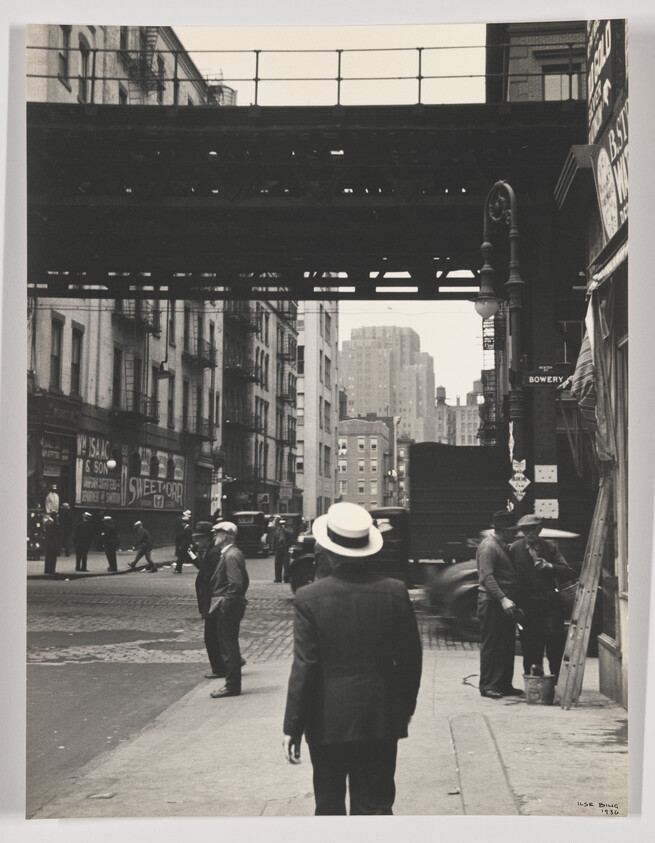 Man in a white hat walks toward elevated train tracks on a busy city street.