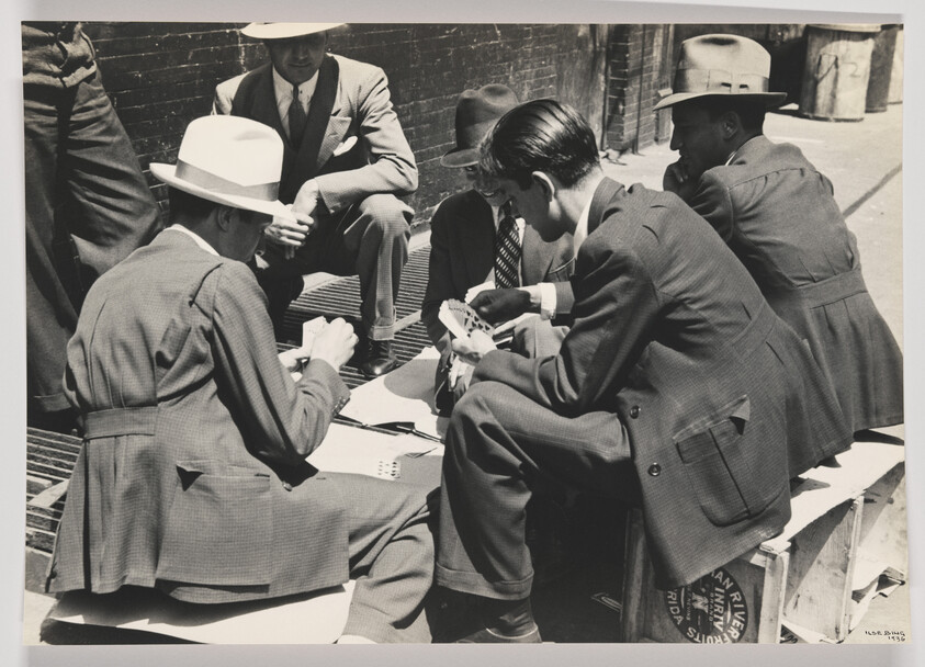 Young men in suits and hats sit on crates playing cards on a city sidewalk.
