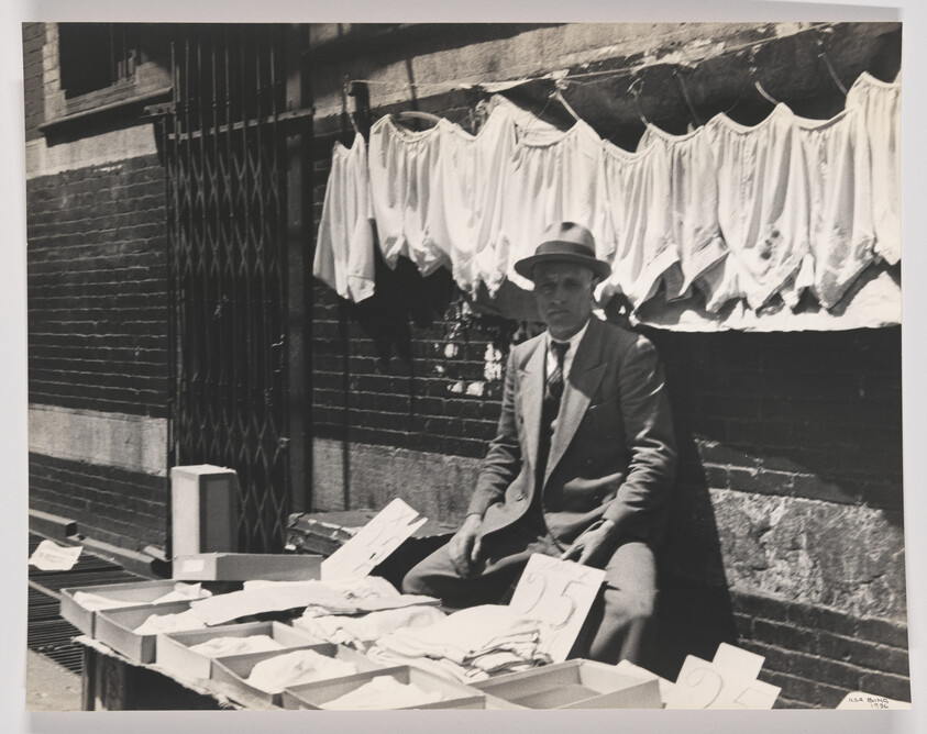 A man in a suit and hat sells undershirts from boxes on a sidewalk market stall.