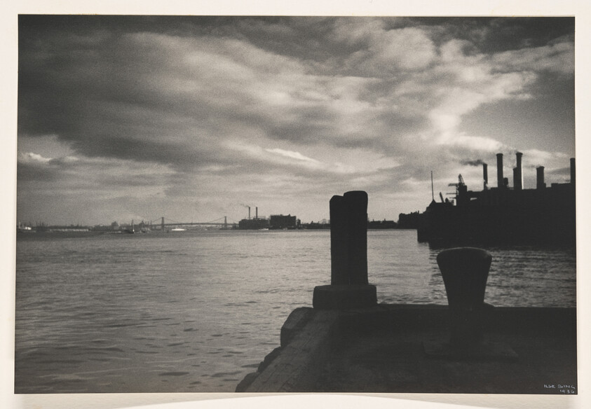 A riverside pier with bollards in foreground and industrial smokestacks silhouetted across the water.