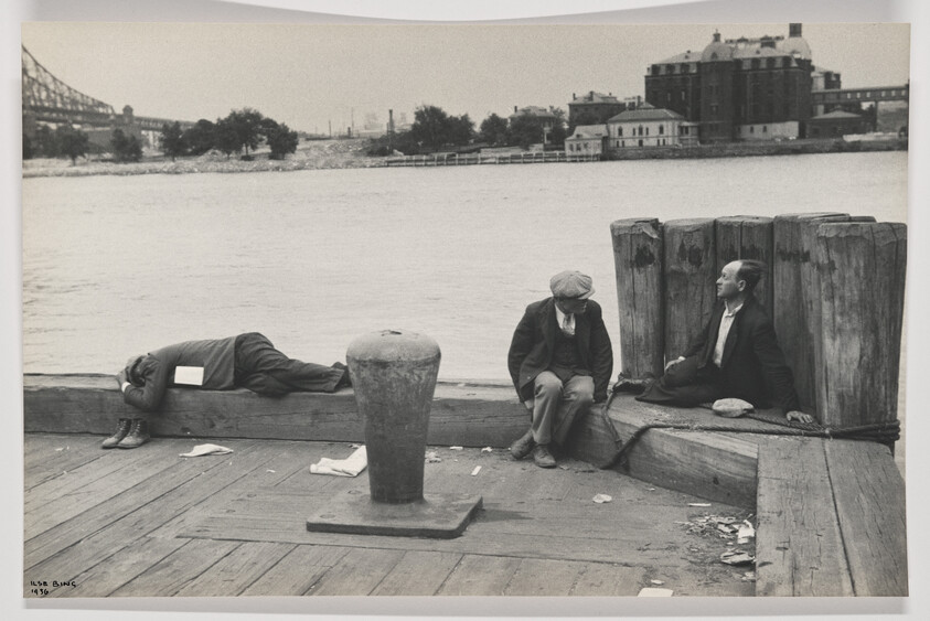 Three men rest on a wooden dock by the river, one lying down and two sitting near pilings.