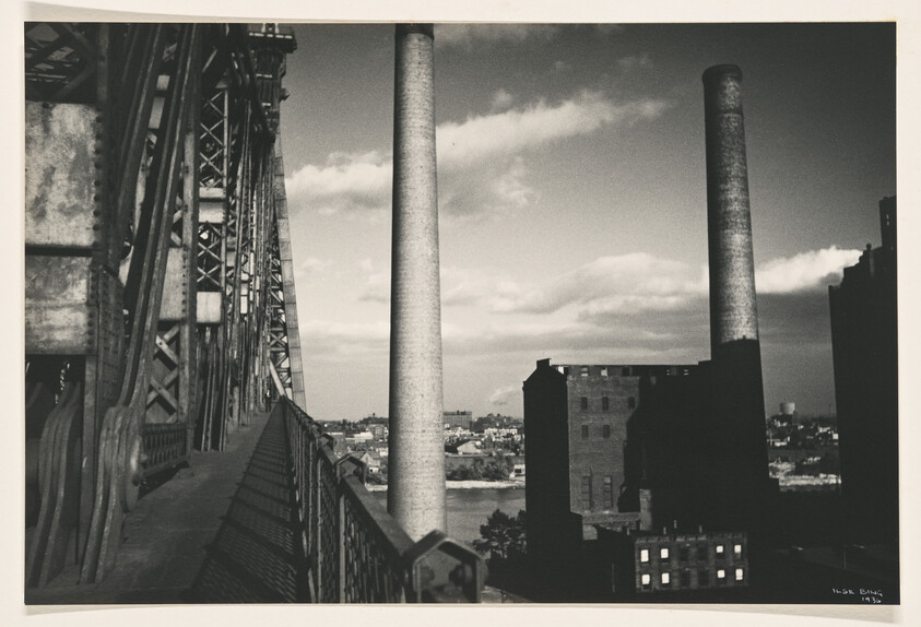 Elevated bridge walkway leading past steel trusses toward tall factory smokestacks and river beyond.