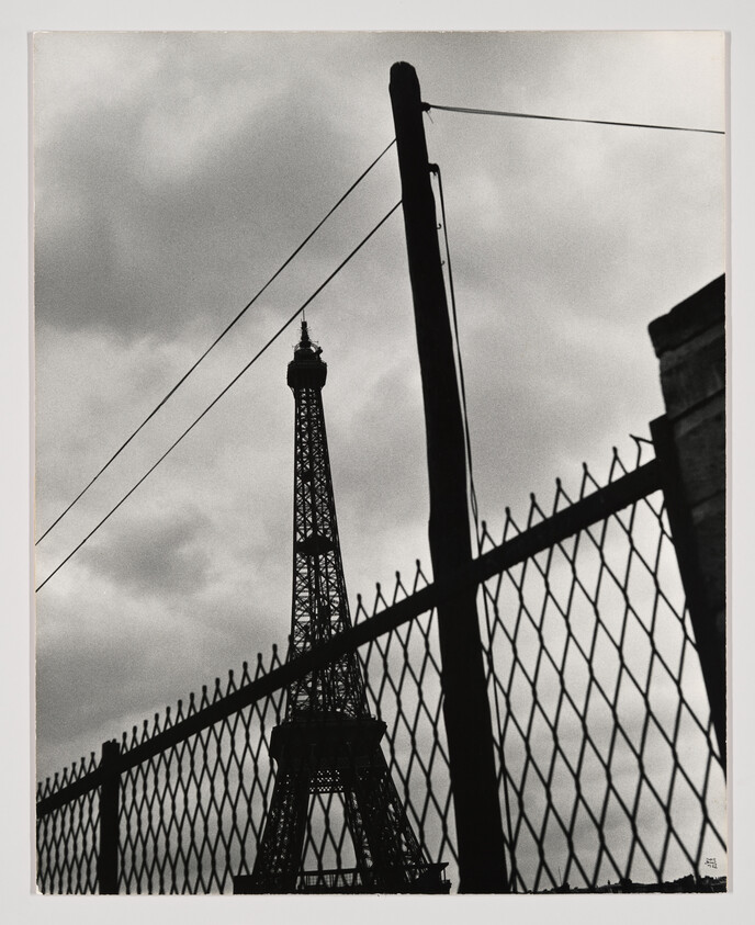 The Eiffel Tower rises behind a chain-link fence and leaning utility pole under cloudy skies.