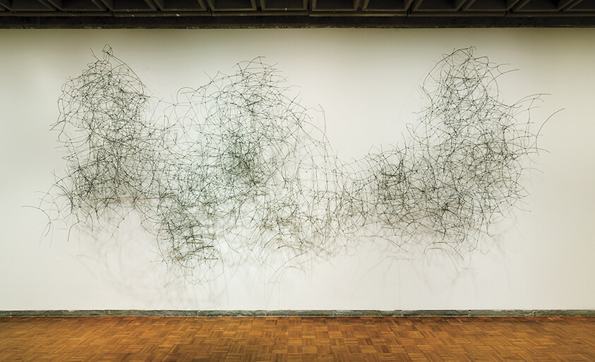 Large tangled wire sculpture mounted on a white gallery wall casting intricate shadows over wooden floor.