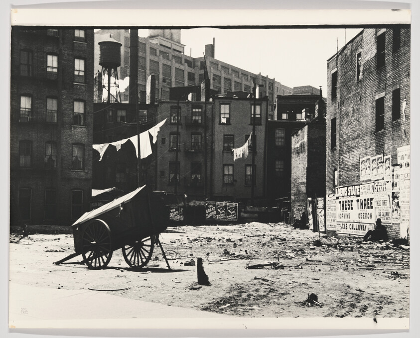 Handcart sits in a vacant urban lot with clotheslines of laundry and old brick buildings behind.