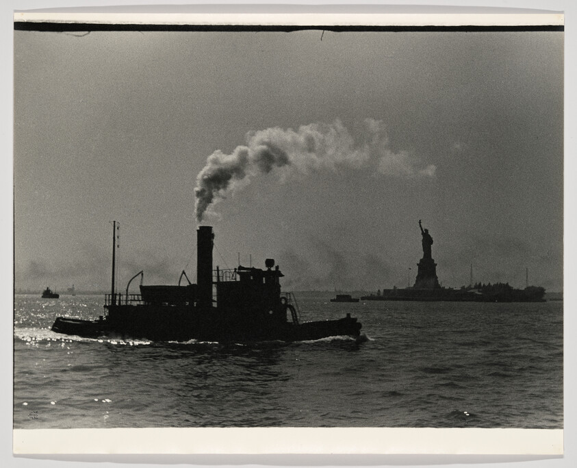 Silhouetted steam tugboat emitting smoke moves across the harbor with the Statue of Liberty in the background.