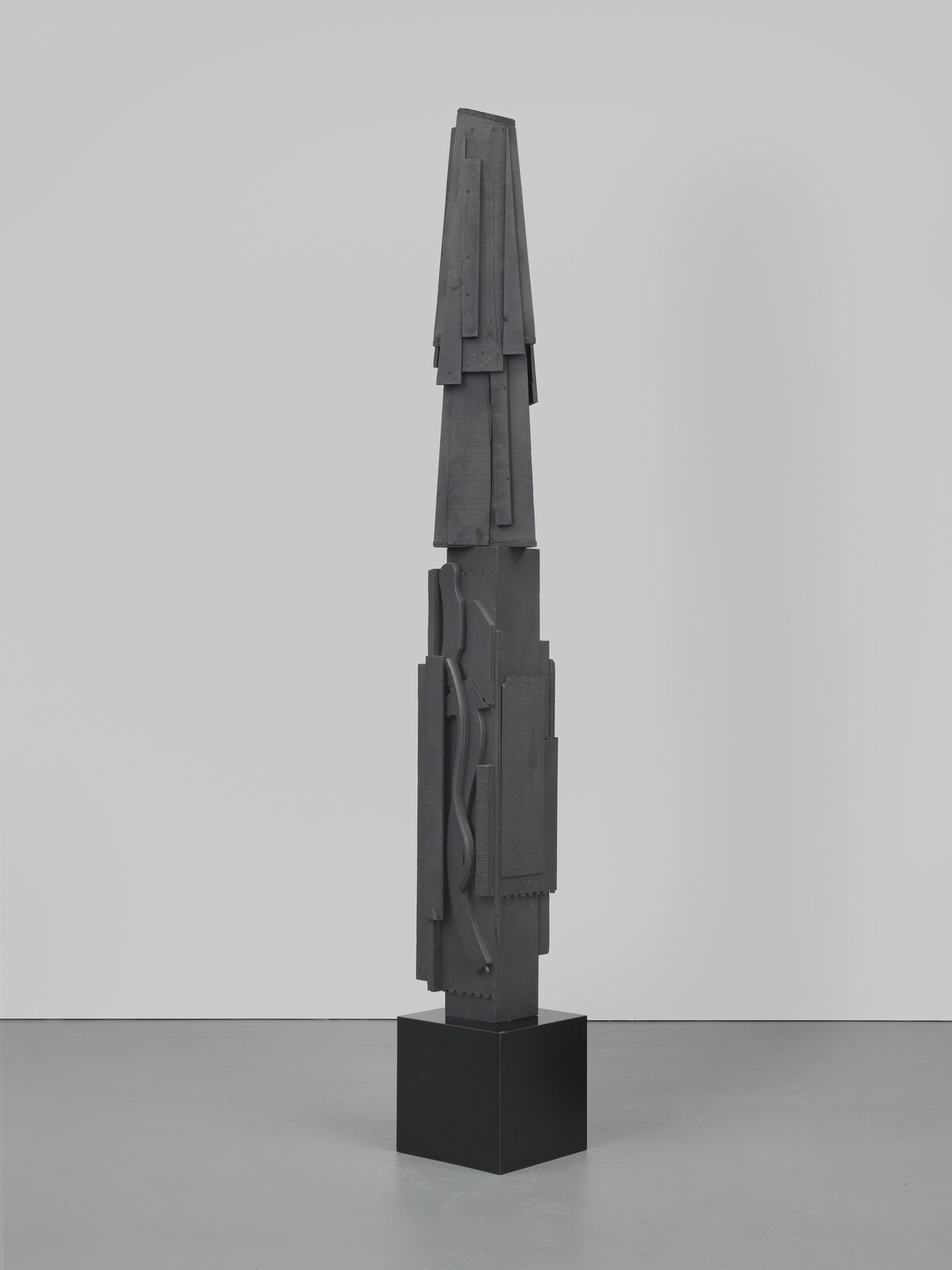 Tall, abstract black sculpture with geometric shapes and textures, standing on a square base.