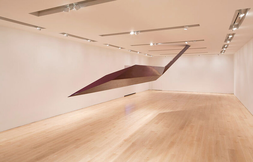 Large, angular sculpture hangs from the ceiling in an empty, well-lit gallery with wooden floors and white walls.