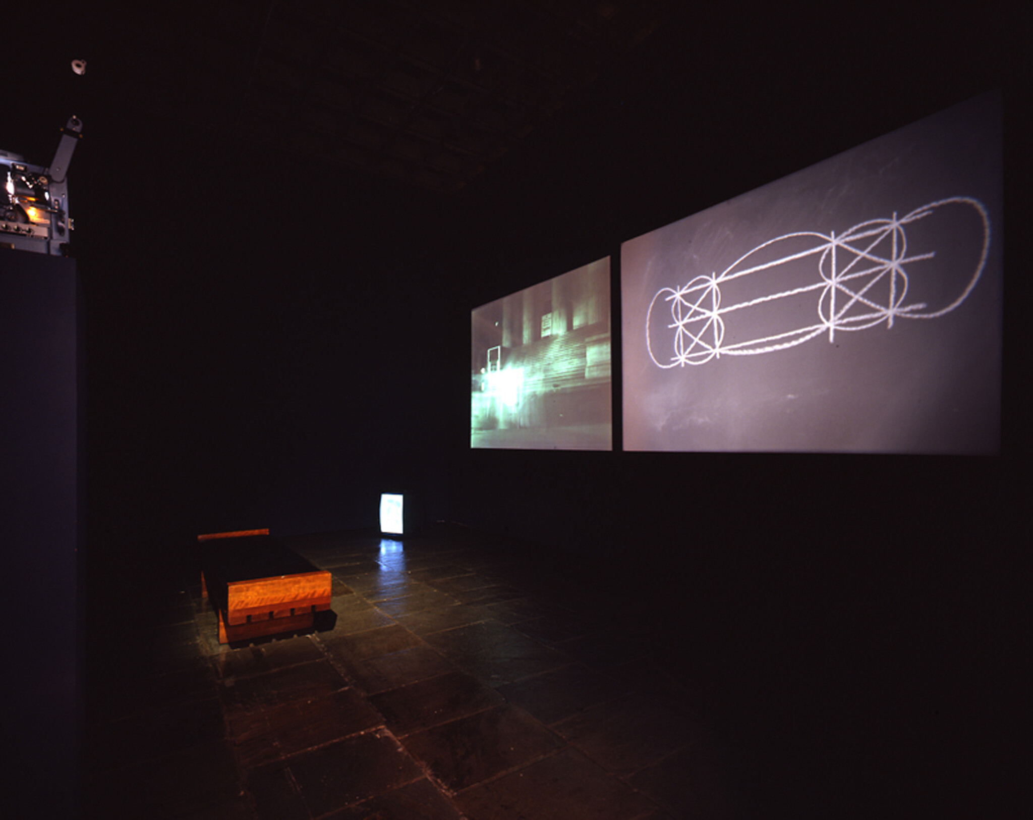 Dark gallery room with two large projected screens showing diagrams and a bench with small TV.