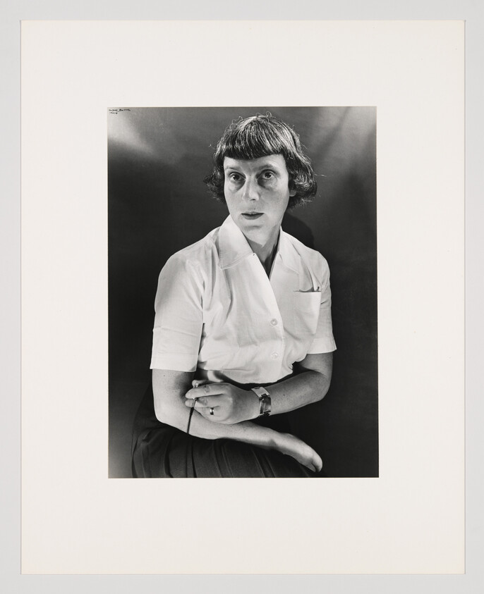 A woman with short hair sits wearing a white blouse, holding eyeglasses and looking sideways.