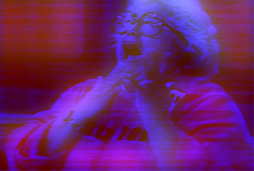 Older woman with glasses wearing a red shirt, hands near her mouth, appears to be shouting or cheering.