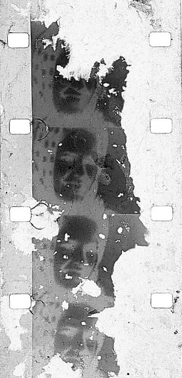 Vertical strip of old film with torn emulsion, dark blotches, and visible sprocket holes.