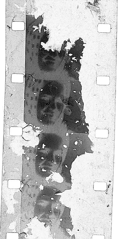 Vertical strip of old film with torn emulsion, dark blotches, and visible sprocket holes.