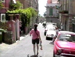 A person wearing pink walks down a narrow street with parked cars and old buildings on both sides.