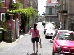 A woman in a pink shirt walks down a narrow residential street carrying shopping bags.