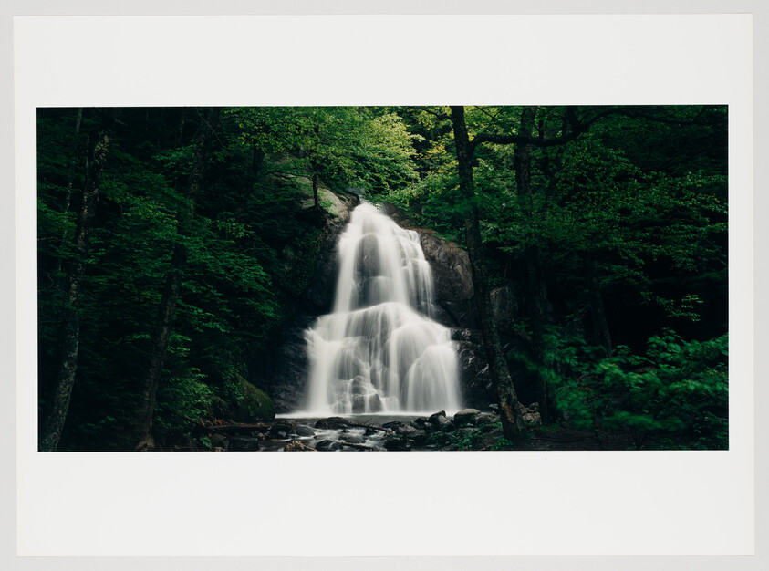 A serene waterfall cascades over a rocky cliff surrounded by lush green foliage in a forest setting, with the water appearing smooth and misty due to a long exposure photograph.